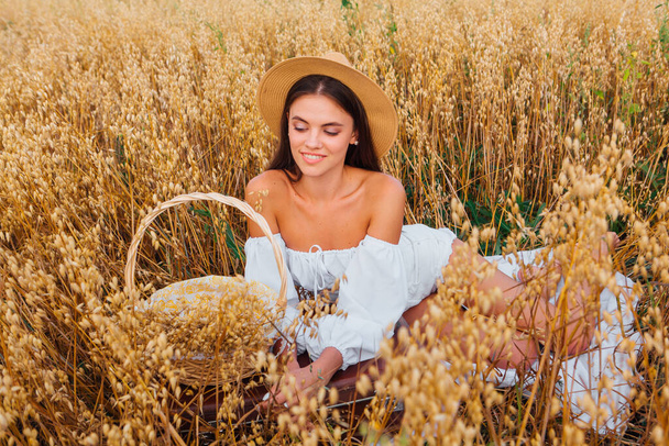 Rural Countryside Scene. Young beautiful woman with long hair dressed in white blouse and straw hat sitting at golden oat field near the basket with ears of oats. - Фото, изображение