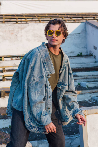 Fashion portrait of a tall handsome man dressed in jeans jacket ans sunglasses on the crushed tribune of the old stadium during sunset - Foto, afbeelding