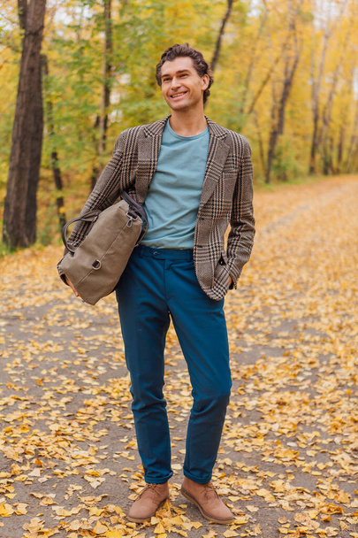 Tall handsome man dressed in a brown jacket walking with a bag on the autumn alley - Foto, Bild
