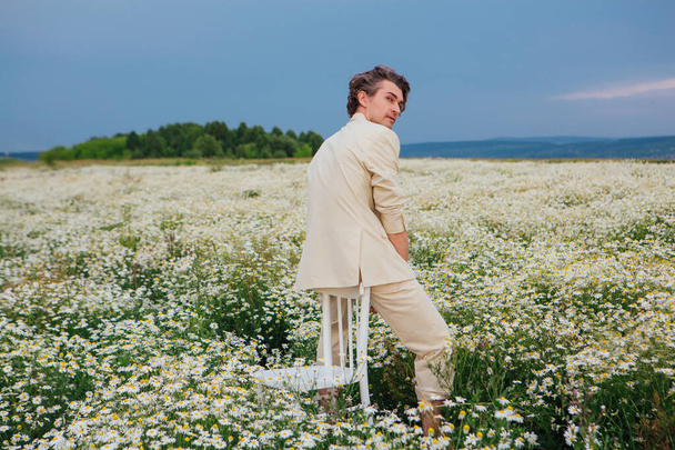 Tall handsome man dressed in a white suit on naked body sitting on a back of a chair in camomile flowers field - Фото, изображение