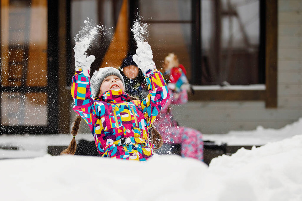 Зимние развлечения, отдых и семейный отдых. Девушка с косичками бросает снег в сугроб перед окном дома и семьи. Выборочный фокус, размытый фон. - Фото, изображение