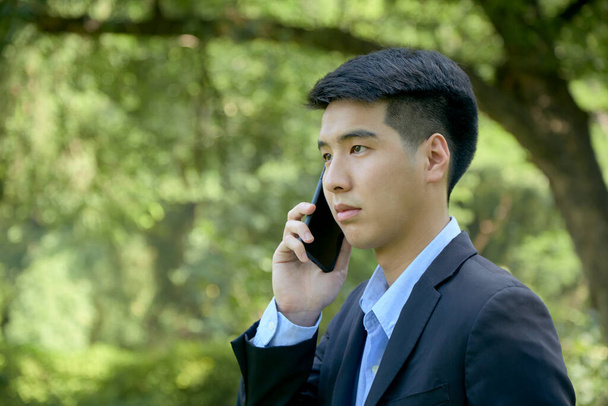 Mladý asijský podnikatel procházky v parku pomocí chytrého telefonu. - Fotografie, Obrázek