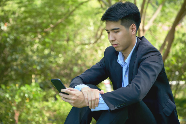 Giovane uomo d'affari asiatico a piedi nel parco utilizzando uno smart phone. - Foto, immagini