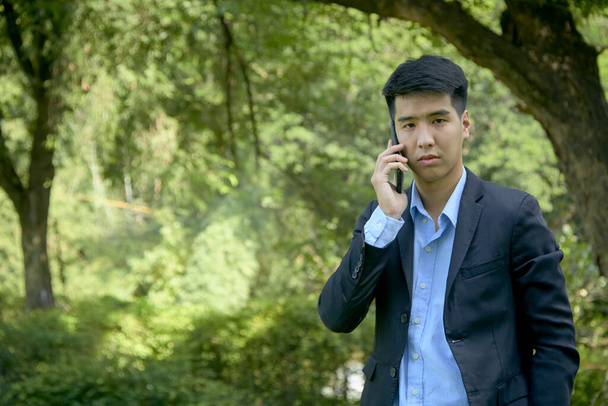 Młody azjatycki biznesmen spacerujący po parku za pomocą smartfona. - Zdjęcie, obraz