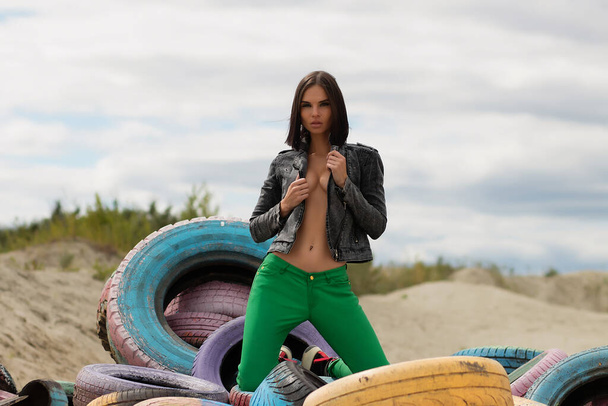 Sensual mujer delgada en chaqueta de moda y pantalones verdes tocando el pelo y mirando a la cámara mientras se arrodilla sobre neumáticos pintados en la playa - Foto, Imagen