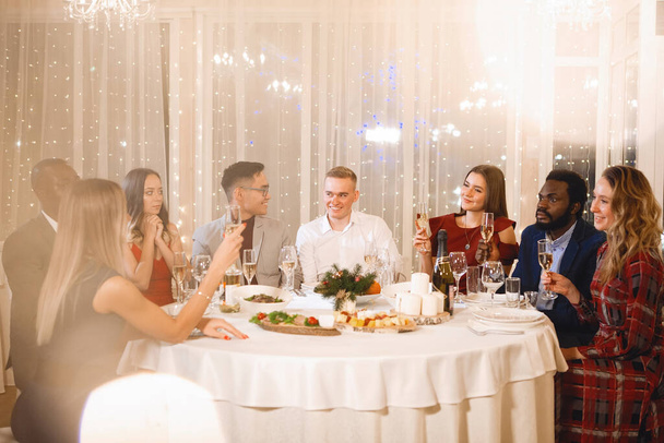 Společnost nadnárodních lidí sedí u slavnostního stolu s sklenicemi šampaňského, usmívá se a mluví v restauraci. Selektivní zaměření, rozmazaná světla - Fotografie, Obrázek