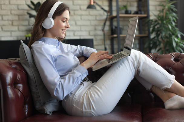 Smiling young woman with headphones and laptop on the sofa - Foto, afbeelding