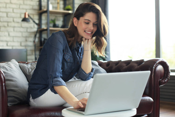 Smiling young woman sitting on sofa with laptop computer and chating with friends - Foto, imagen