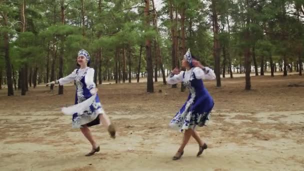 Dos hermosas mujeres en traje popular tradicional ruso azul con pañuelo están bailando en el bosque - Imágenes, Vídeo