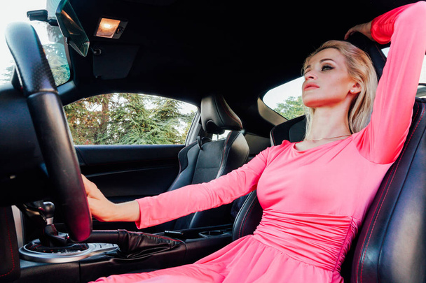 Όμορφη γυναίκα ξανθιά οδηγός σε ροζ φόρεμα πίσω από το τιμόνι ενός αυτοκινήτου - Φωτογραφία, εικόνα