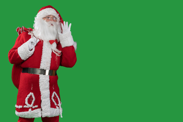 santa claus op een groene achtergrond met een grote rode zak staat met zijn hand op zijn mond en schreeuwt. Begrepen, ruimte. Kerst- en nieuwjaarsconcept - Foto, afbeelding