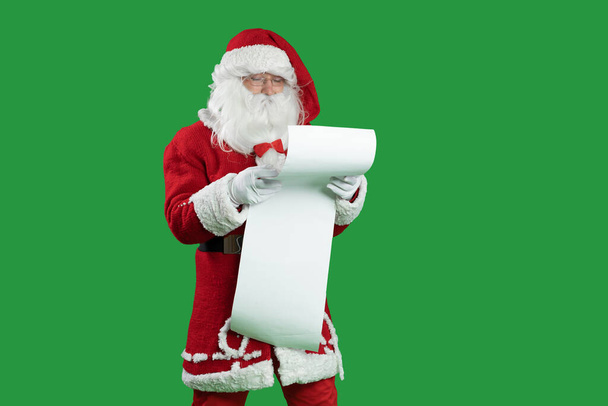 Santa Claus na zeleném pozadí obsahuje seznam dětí, které si jej pečlivě přečtou. Překvapený obličej. Kopírovat prostor. - Fotografie, Obrázek