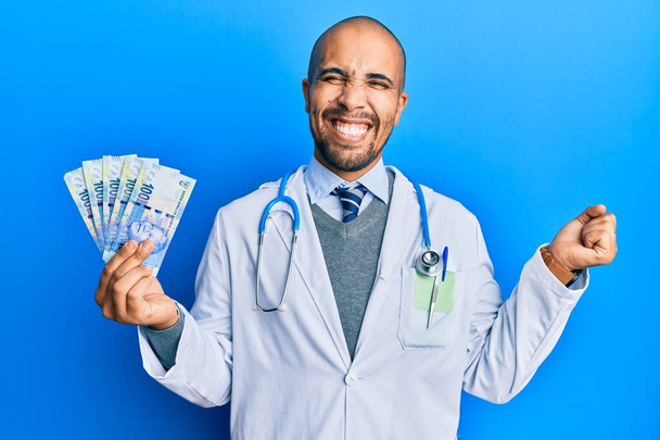 médecin adulte hispanique homme portant l'uniforme médical tenant 100 rand sud-africain criant fier, célébrant la victoire et le succès très excité avec le bras levé  - Photo, image
