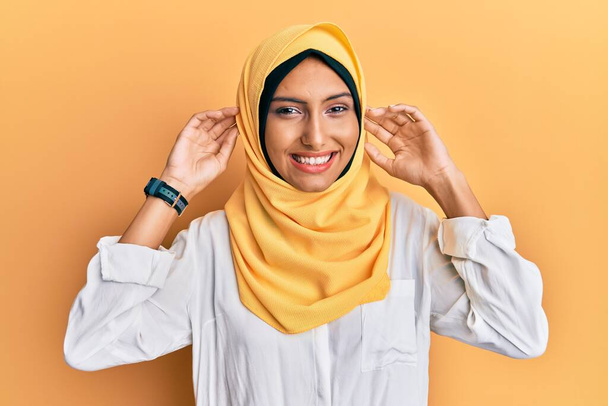 Молода брюнетка арабська жінка в традиційному ісламському шарфі хіджаб посміхається тягнути вуха пальцями, смішний жест. проблема прослуховування
  - Фото, зображення