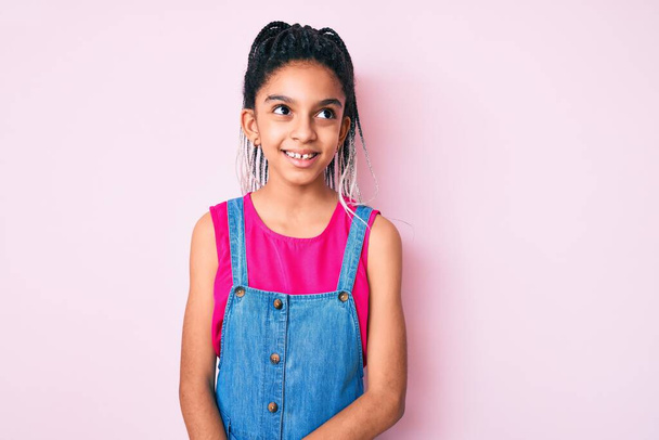 Jeune fille afro-américaine enfant avec des tresses portant des vêtements décontractés sur fond rose regardant de côté avec le sourire sur le visage, expression naturelle. riant confiant.  - Photo, image
