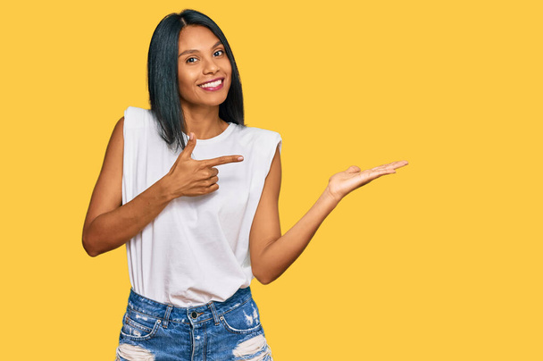 Junge afrikanisch-amerikanische Frau in lässiger Kleidung überrascht und lächelt in die Kamera, während sie mit der Hand präsentiert und mit dem Finger zeigt.  - Foto, Bild