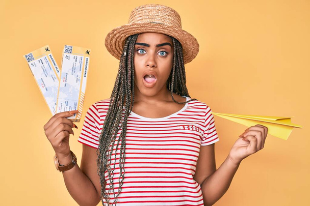 Mujer afroamericana joven con trenzas sosteniendo avión de papel y tarjeta de embarque en la cara de choque, mirando escéptico y sarcástico, sorprendido con la boca abierta  - Foto, imagen