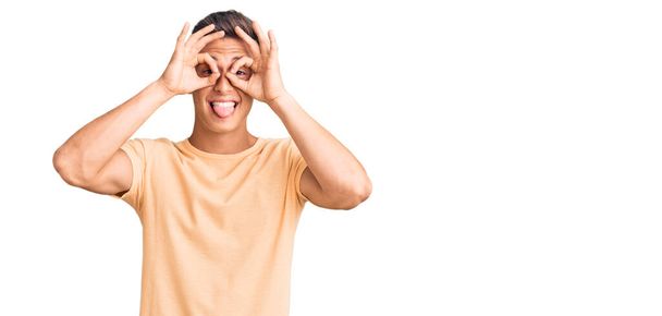 Young handsome man wearing casual clothes doing ok gesture like binoculars sticking tongue out, eyes looking through fingers. crazy expression.  - Foto, Bild