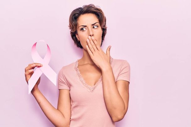 Mladá krásná žena drží růžovou rakovinu stužku zakrývající ústa rukou, šokovaná a vystrašená z omylu. překvapený výraz  - Fotografie, Obrázek