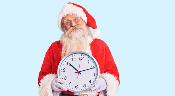 Gri saçlı, uzun sakallı, Noel Baba kostümü giyen yaşlı bir adam saat tutuyor. Pozitif ve mutlu görünüyor. Kendine güvenen bir gülümsemeyle dişlerini gösteriyor.  - Fotoğraf, Görsel