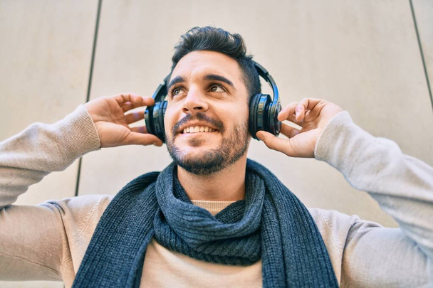 Genç İspanyol gülümseyen adam şehirde kulaklık takarak müzik dinlemekten mutlu oluyor.. - Fotoğraf, Görsel