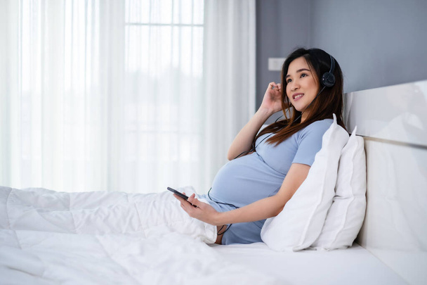 pregnant woman using smartphone to listening music through headphones on a bed - Foto, afbeelding