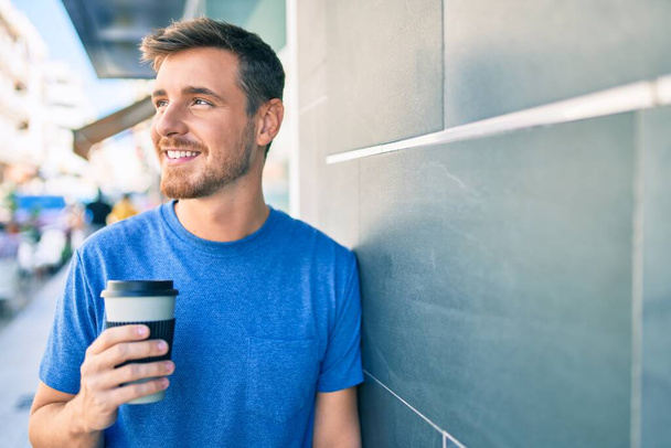 若い白人男性がスマートフォンを使って街でコーヒーを飲んでいます. - 写真・画像