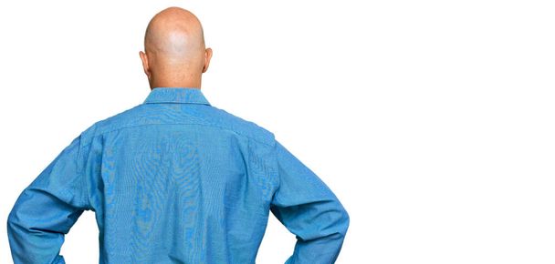 Homme chauve d'âge moyen portant des vêtements décontractés debout vers l'arrière regardant loin avec les bras sur le corps  - Photo, image