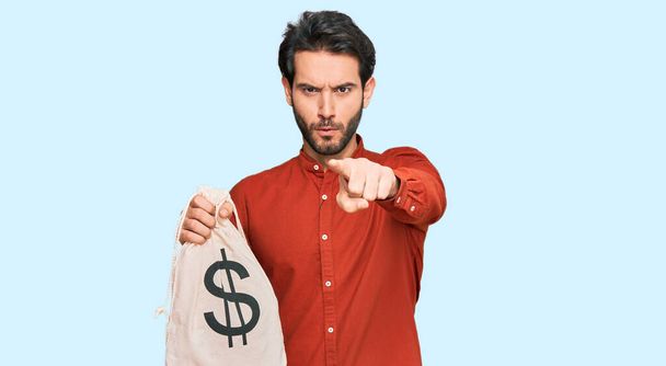 Jeune homme hispanique tenant sac dollars pointant du doigt vers la caméra et à vous, geste confiant regardant sérieux  - Photo, image