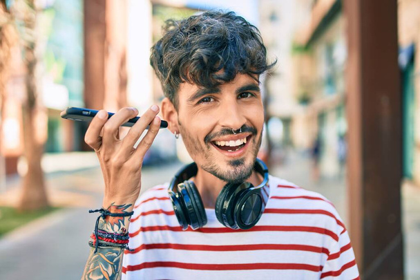 Joven hombre hispano usando auriculares y hablando en el teléfono inteligente en la calle de la ciudad - Foto, Imagen