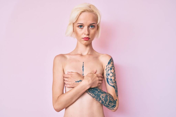 Jonge blonde vrouw met tatoeage staat shirtloos ontspannen met serieuze uitdrukking op het gezicht. eenvoudig en natuurlijk kijkend naar de camera.  - Foto, afbeelding