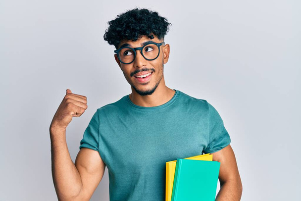Young arab handsome man wearing glasses and holding books pointing thumb up to the side smiling happy with open mouth  - 写真・画像