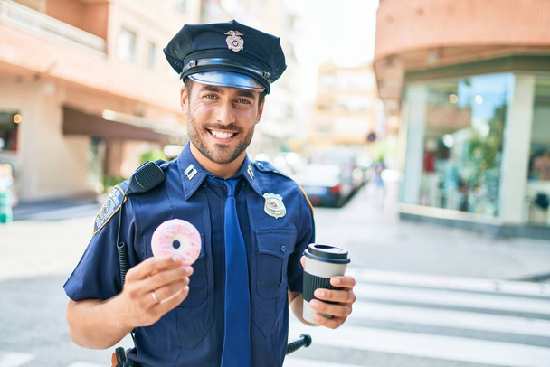Mladý pohledný hispánský policista v policejní uniformě se šťastně usmívá. Jíst koblihy a pití šálek odnést kávu na ulici města. - Fotografie, Obrázek