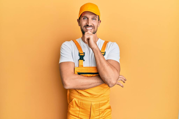 Young handsome man wearing handyman uniform over yellow background looking confident at the camera smiling with crossed arms and hand raised on chin. thinking positive.  - Foto, afbeelding