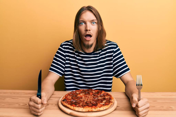 Schöner kaukasischer Mann mit langen Haaren, der leckere Pfefferoni-Pizza isst, ängstlich und schockiert mit Überraschung und erstauntem Gesichtsausdruck, Angst und aufgeregtem Gesicht.  - Foto, Bild