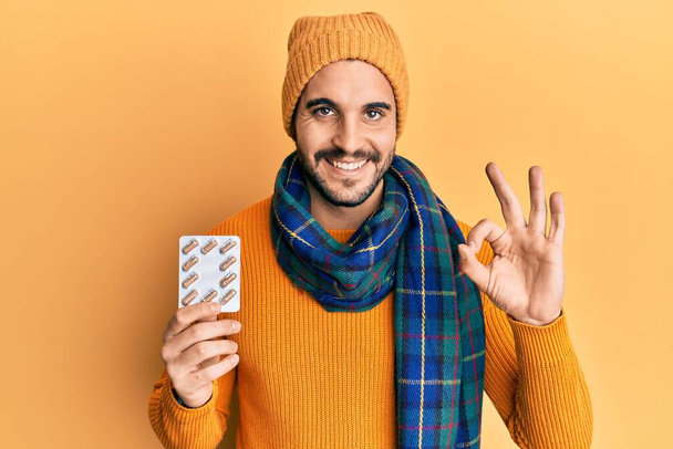 Joven hombre hispano con suéter de lana y bufanda de invierno sosteniendo píldoras haciendo signo bien con los dedos, sonriendo gesto amistoso excelente símbolo  - Foto, imagen