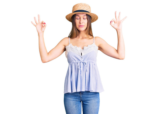 Красива брюнетка молода жінка в літньому капелюсі розслабляється і посміхається з закритими очима, роблячи медитаційний жест пальцями. йога концепція
.  - Фото, зображення
