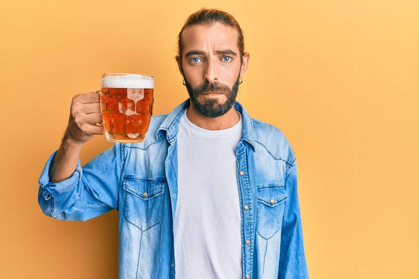 Atraktivní muž s dlouhými vlasy a vousy pije pintu piva myšlení postoj a střízlivý výraz vypadá sebevědomě  - Fotografie, Obrázek