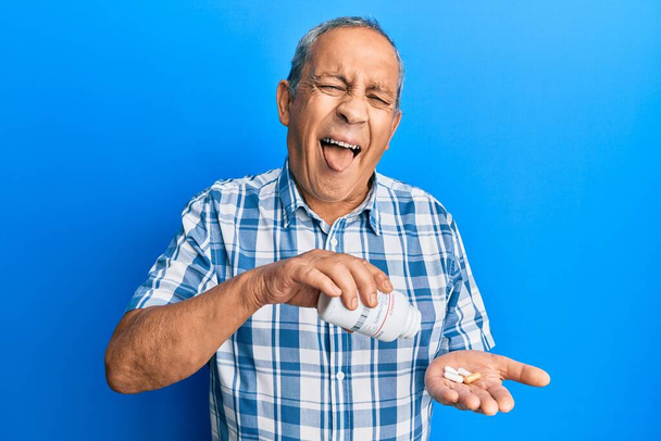Όμορφος ηλικιωμένος άνδρας με γκρίζα μαλλιά κρατώντας χάπια στο πρόσωπο σοκ, δείχνοντας σκεπτικός και σαρκαστικός, έκπληκτος με ανοιχτό στόμα  - Φωτογραφία, εικόνα