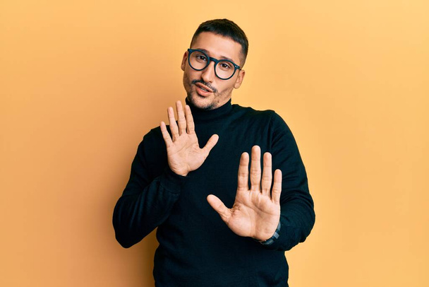 Handsome man with tattoos wearing turtleneck sweater and glasses moving away hands palms showing refusal and denial with afraid and disgusting expression. stop and forbidden.  - Photo, Image