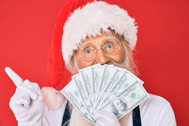 Старий старший чоловік з сірим волоссям і довгою бородою в костюмі Санти Клауса тримає долари посміхаючись щасливо вказуючи рукою і пальцем на бік
  - Фото, зображення