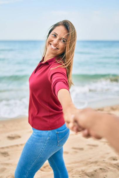 Piękna kobieta w średnim wieku trzymająca się za ręce z mężem na plaży, prowadząca szczęśliwą drogę uśmiechnięta w miłości - Zdjęcie, obraz