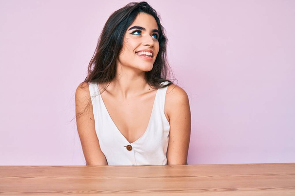 Brunette teenager girl wearing casual clothes sitting on the table looking away to side with smile on face, natural expression. laughing confident.  - Photo, Image