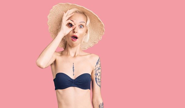 Jonge blonde vrouw met tatoeage in bikini en zomerhoed doet ok gebaar geschokt met een verrast gezicht, oog kijkend door vingers. ongelovige uitdrukking.  - Foto, afbeelding