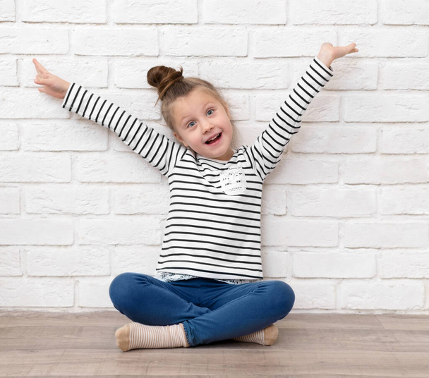 Happy little Caucasian girl 5-6 years old sitting on the floor against a white brick wall with her arms outstretched. The concept of a happy childhood, activities and games at home during the pandemic - Foto, Imagem