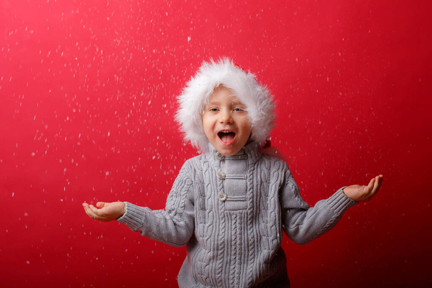 um menino em um chapéu de Papai Noel pega neve em um fundo vermelho - Foto, Imagem