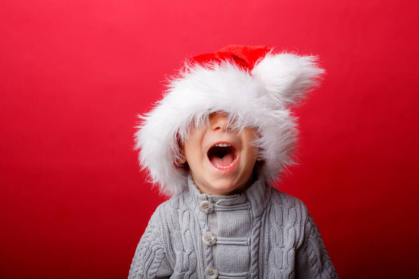 赤いサンタの帽子の子供の肖像画、クリスマスの夜に面白い子供を笑顔。子供は帽子で目を覆いました - 写真・画像