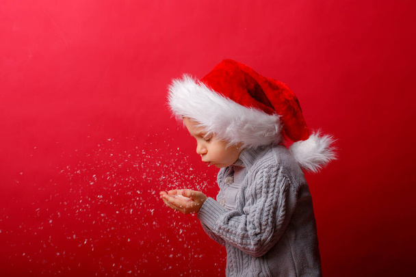 a boy in a Santa Claus hat blows snow on a red background - Photo, Image