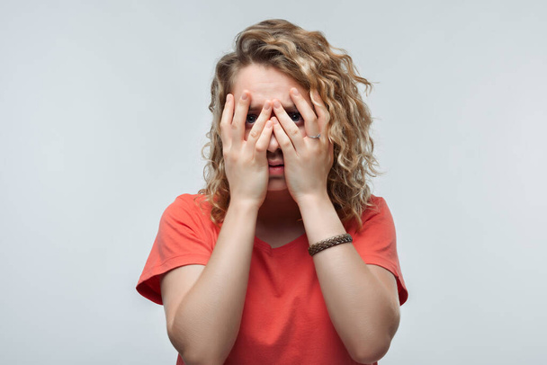 Portrait of frightened young woman with curly hair in casual t shirt covering her eyes with hands. Human emotions, facial expression concept. Studio shot, white background, isolated - 写真・画像