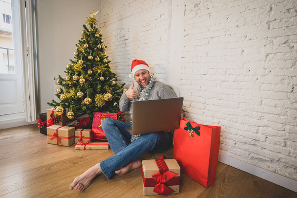 Happy man home alone in lockdown celebrating virtual christmas using laptop video calling family and friends. Virtual holiday gathering online due to coronavirus quarantines and social distancing. - 写真・画像